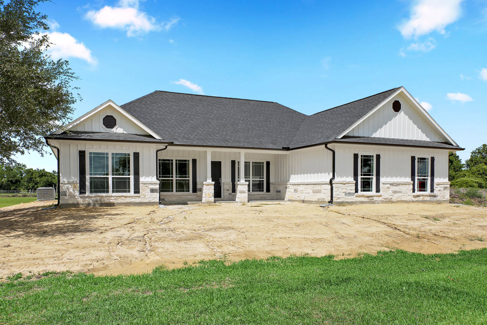 White house with black roof, driveway bordered by green grass, leafy tree near front, black vent on exterior wall, large metal utility box with blue handle visible.