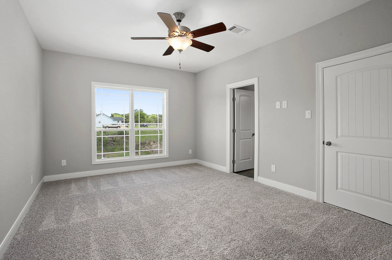 Bedroom with beige carpet, white walls, ceiling fan with light fixture, large window overlooking neighboring house and field, white door with silver handle