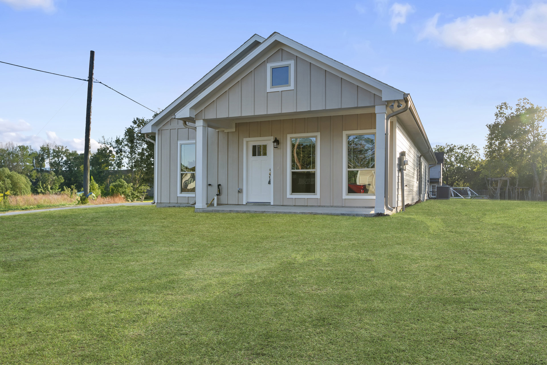 Grey siding house with white door and windows, surrounded by green lawn, small porch, and trees in the background under partly cloudy sky
