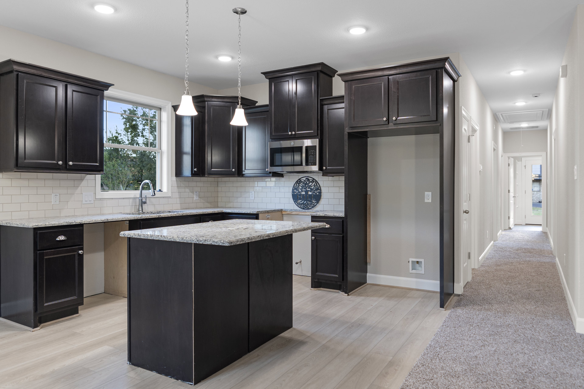 Kitchen with dark wood cabinets, white tile floor, granite-topped island, black cabinet with silver knobs, microwave oven with black door, metal wall sign, and white square box