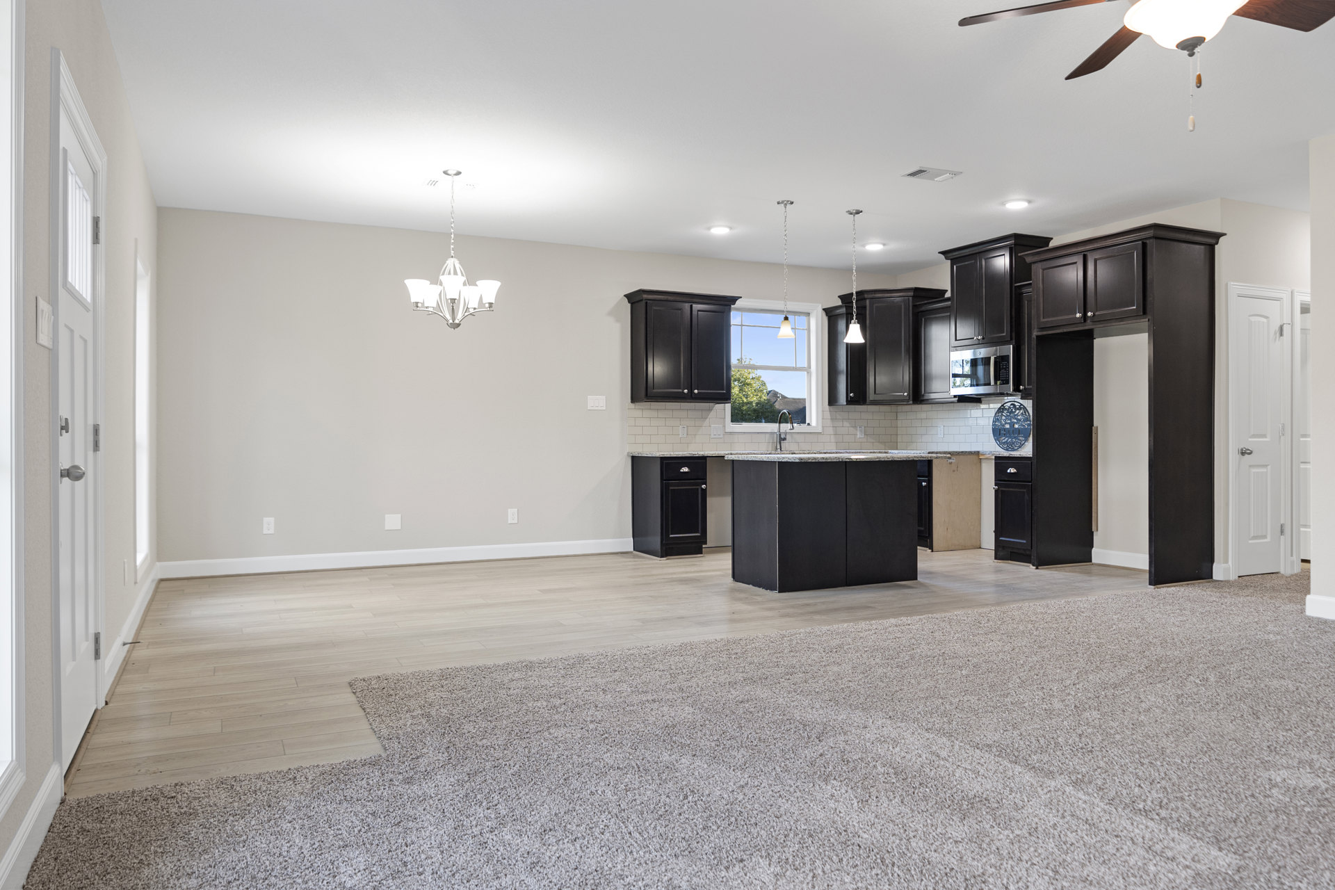 Open-concept kitchen and living room featuring a ceiling fan, tile backsplash with a metal sign, black cabinetry with silver knobs, black rectangular countertop, microwave oven