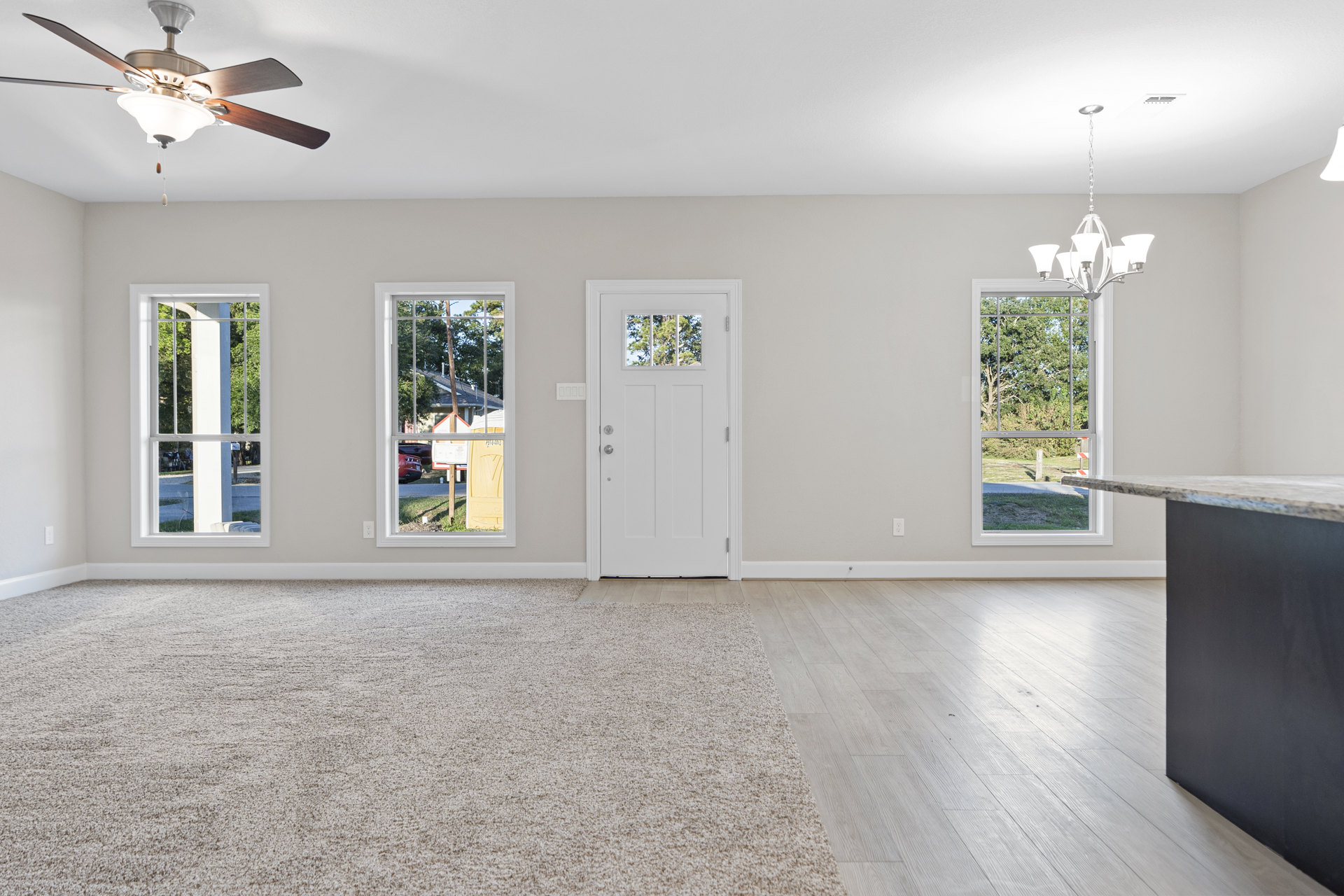 Living room with wood flooring, ceiling fan with light fixture, large windows showing street view, white door with glass insert, neutral walls, minimal decor