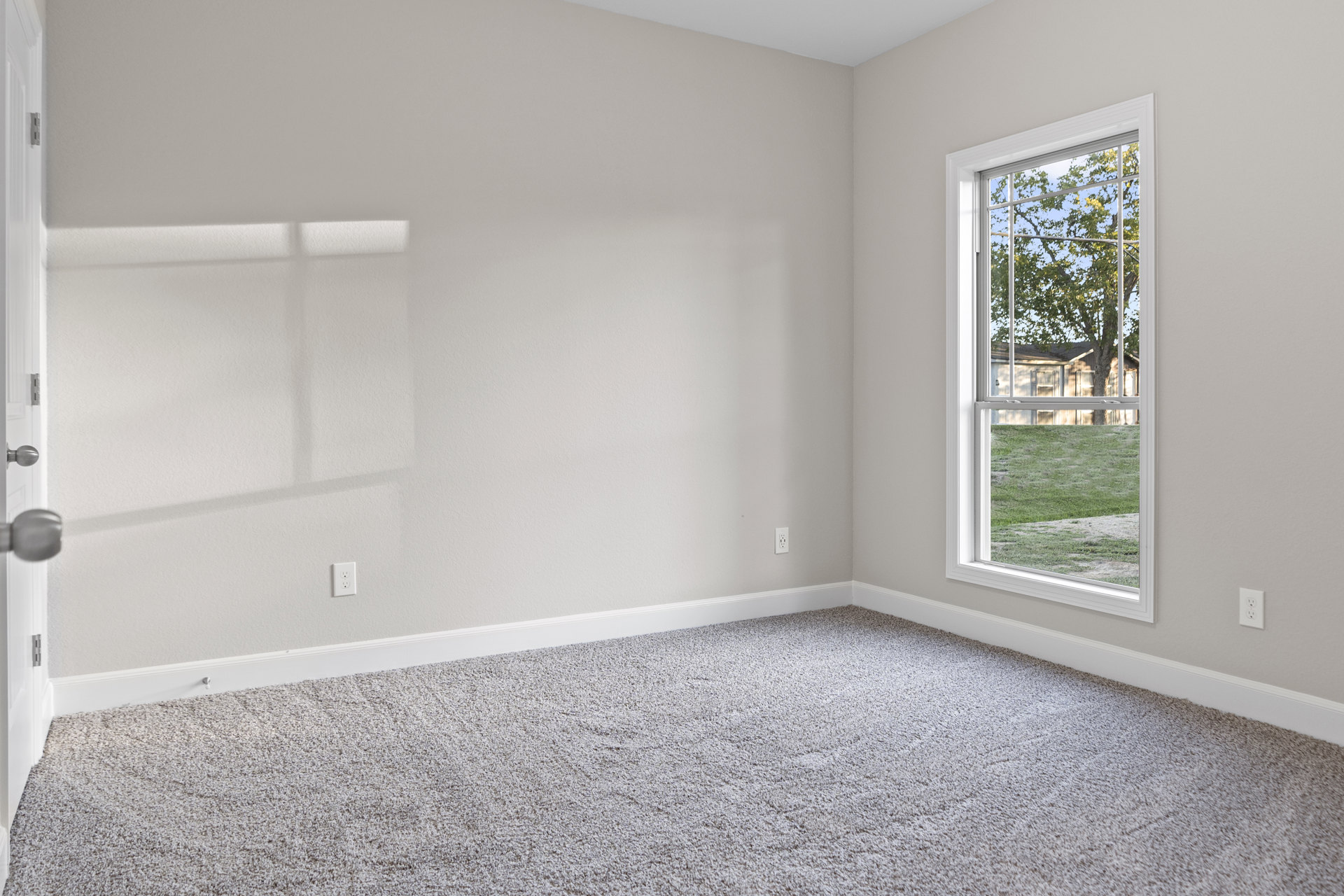 Neutral-toned carpeted room with white walls, large window overlooking green grass and trees, simple ceiling, and a close-up of a brushed metal door knob