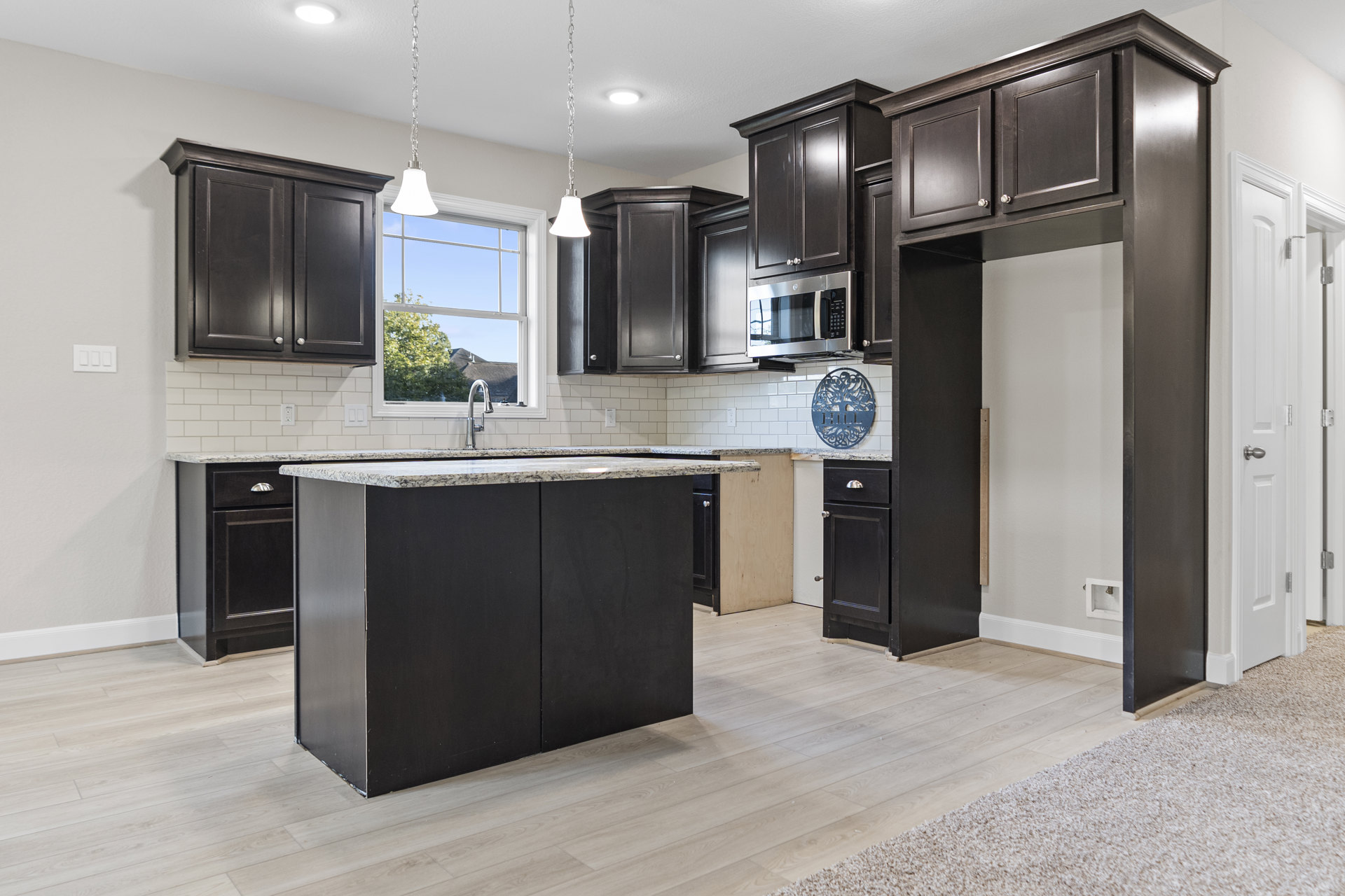 Dark wood kitchen cabinets with silver knobs, black kitchen island topped with marble, white tile floor, microwave oven with black handle, blue sign mounted on tile wall, window