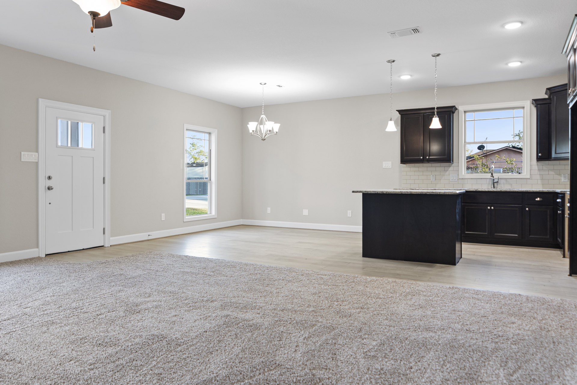 Open-concept living room with patterned rug, adjacent kitchen featuring black countertop, white tile backsplash, white cabinetry, ceiling light fixture, windowed door, and large