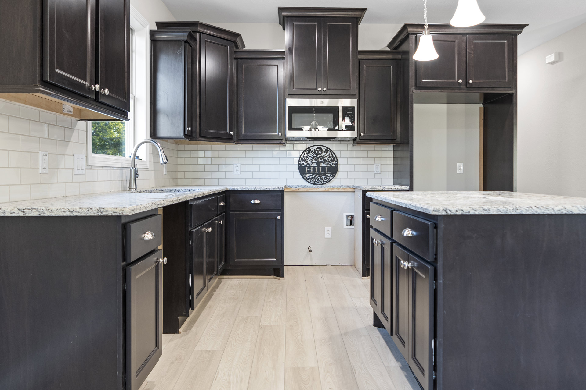 Dark wood kitchen cabinets, white tile flooring, marble-topped island, stainless steel microwave, black door, modern light fixture, black sign on white tile backsplash