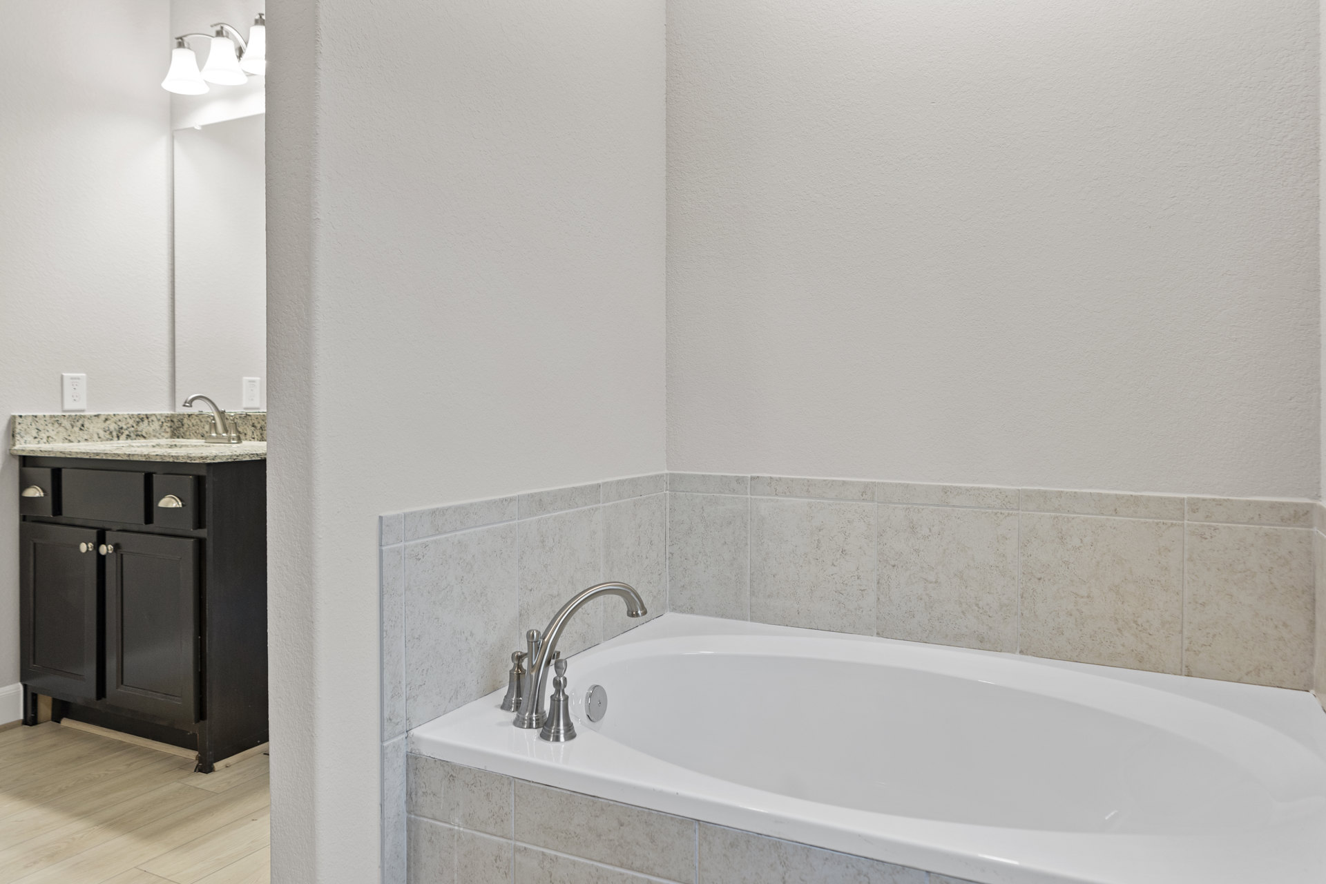 White freestanding bathtub with silver faucet beside black cabinet with silver handles, wood flooring, and three-light fixture in modern bathroom.