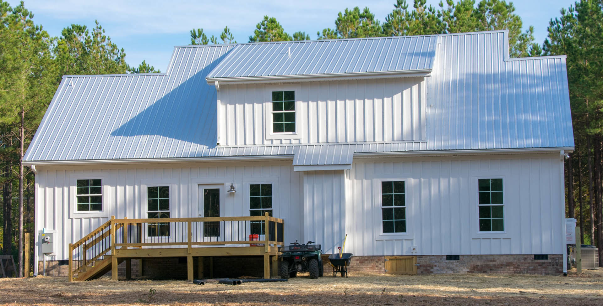 White house with horizontal siding, wooden deck with railing, blue-framed window, wheelbarrow and shovels on deck, green lawn and trees in background.