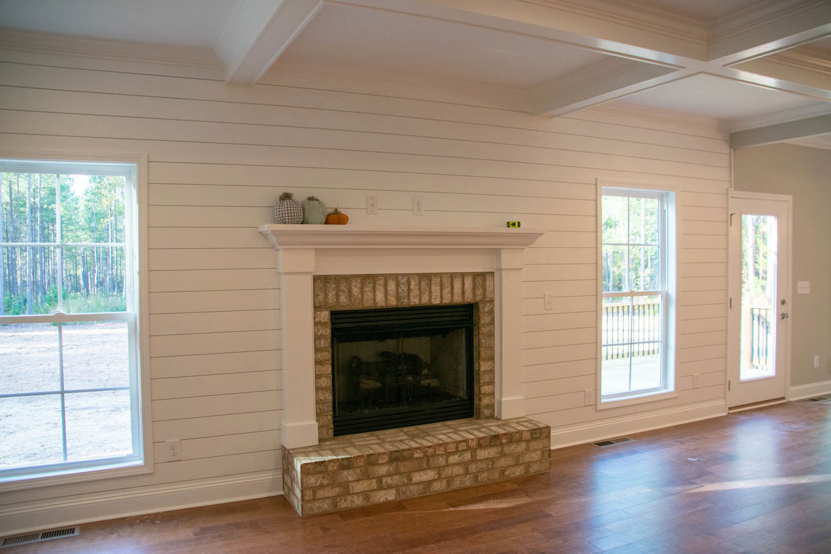 White fireplace with glass door set in a brick wall, white mantle above, large window with forest view and exterior railing, hardwood floors in a cozy den.