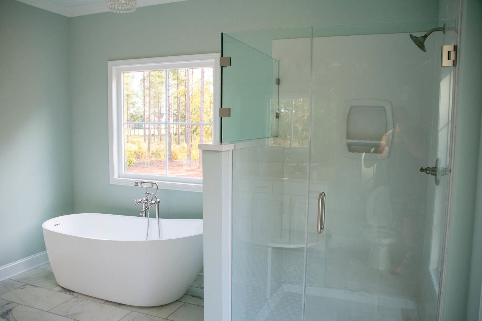 White freestanding bathtub with silver faucet beside glass shower enclosure, large window with view of trees, light tile walls, and modern plumbing fixtures
