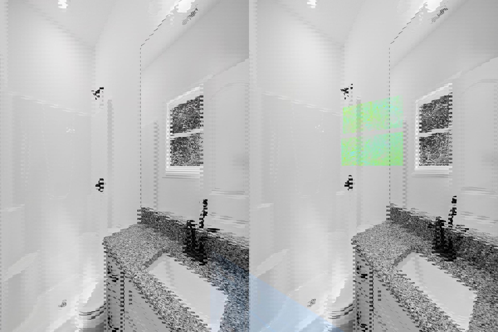 Modern bathroom featuring a white sink with a matte black faucet, black shower head mounted on a white tiled wall, toilet seat, and window overlooking a forest.