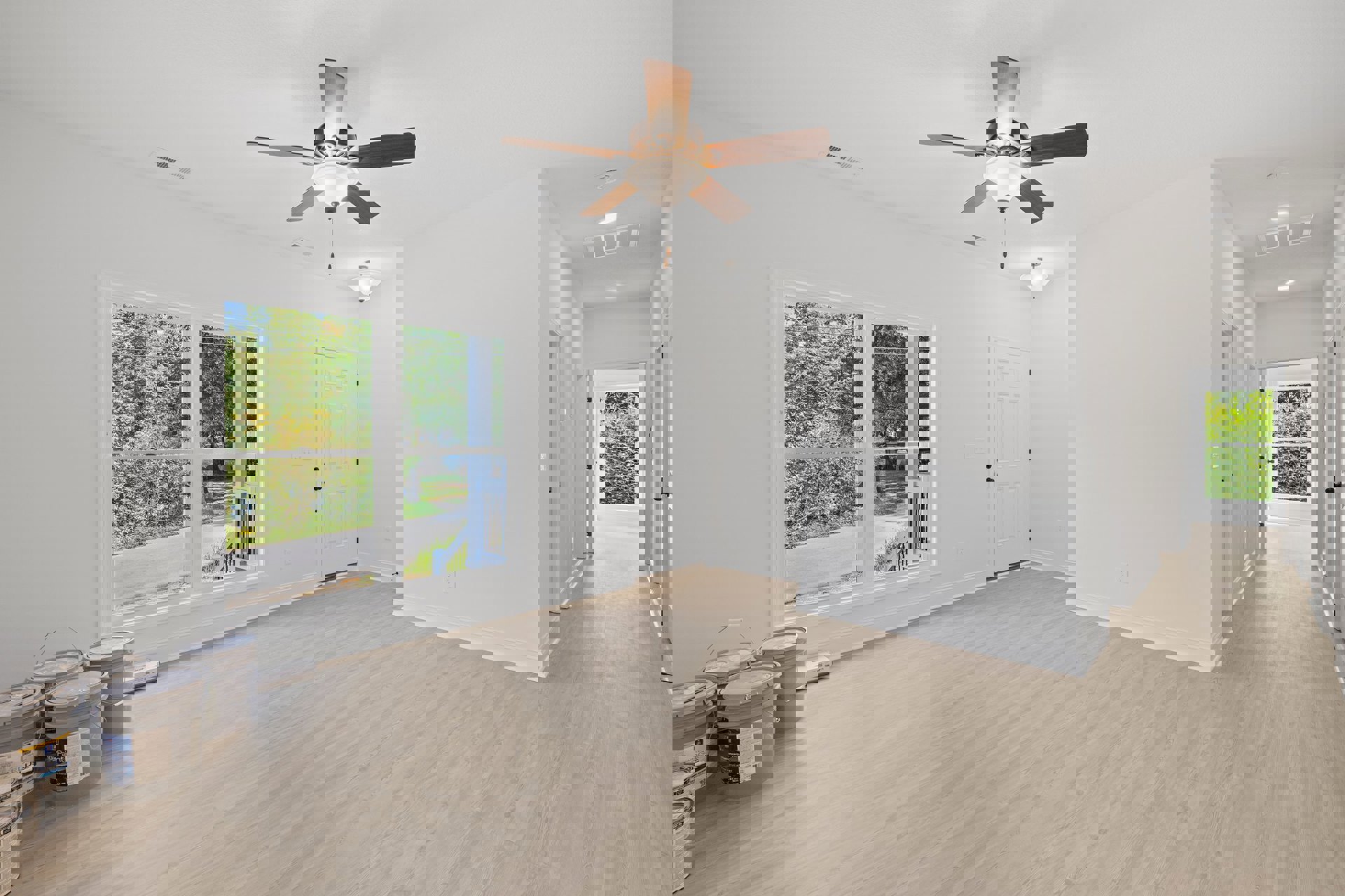 Bedroom with wood laminate flooring, white walls, ceiling fan with light, white-framed window overlooking green plants, white door with black hardware, and paint bucket in the