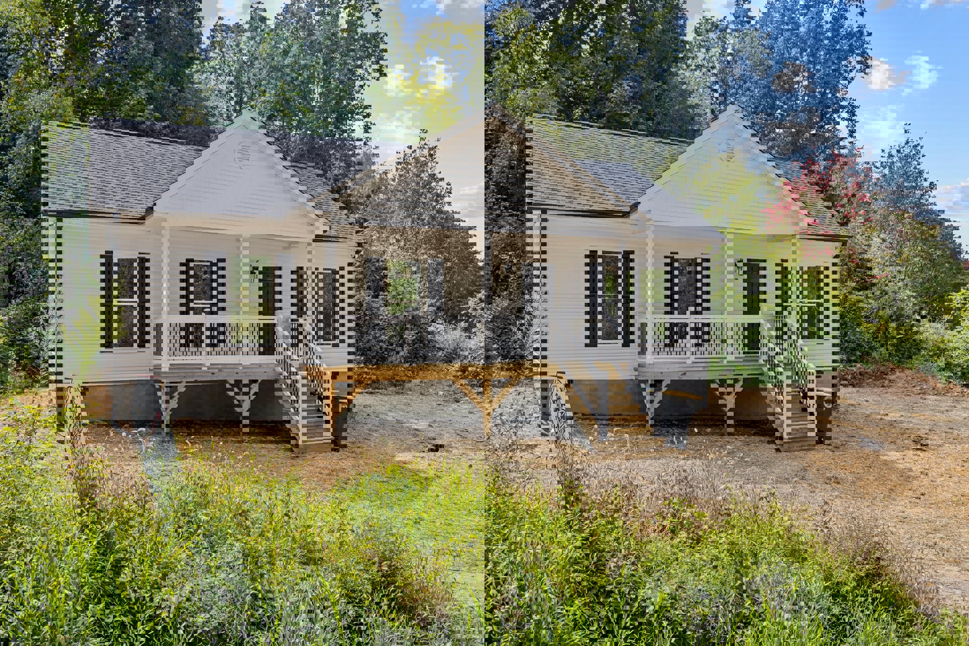 White siding house with two covered porches, wooden deck with white railing, grassy lawn, white-framed windows, and small sign near the foundation.