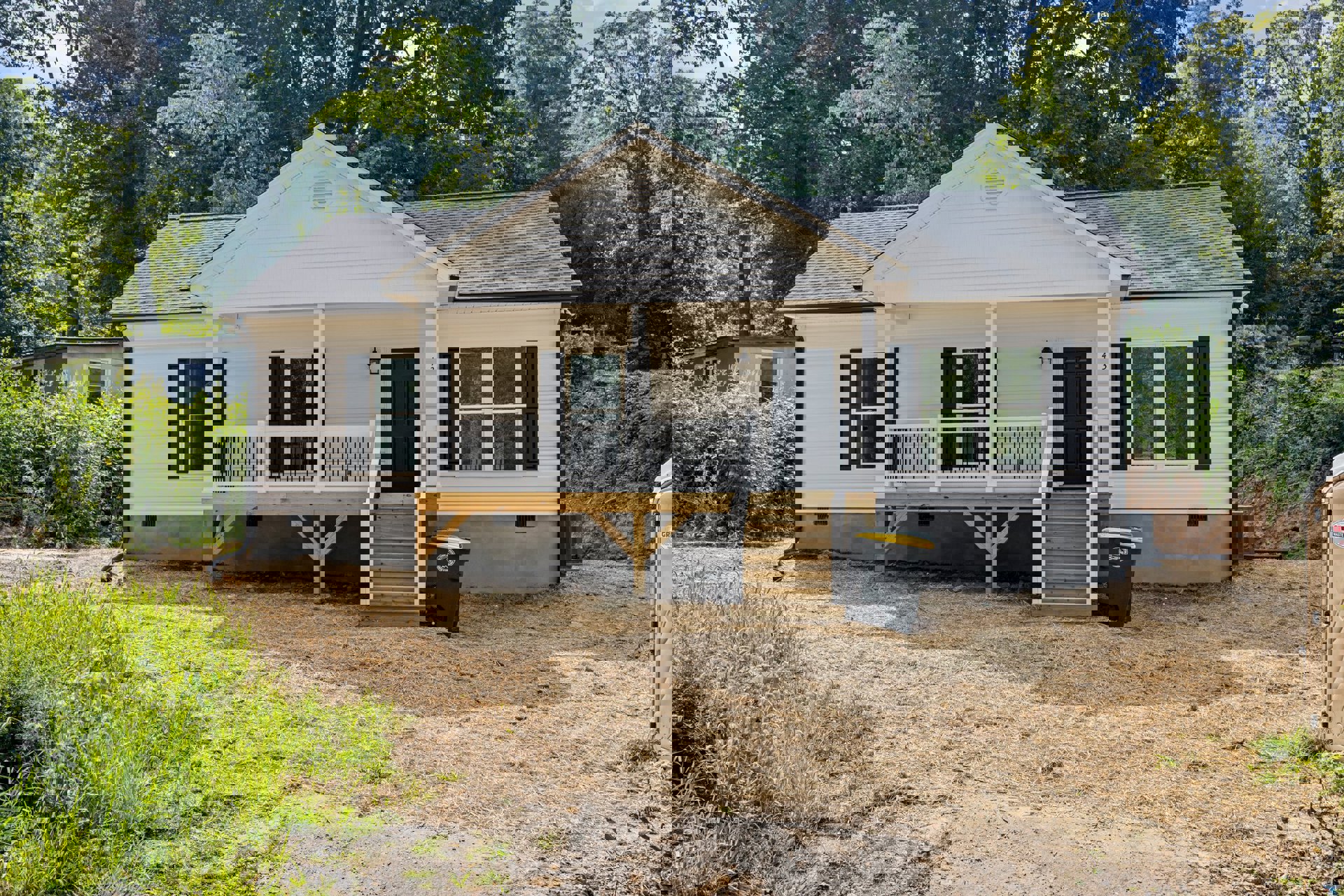 White house with black door, wooden porch deck, black and yellow trash can near vented wall, green grass and dirt yard