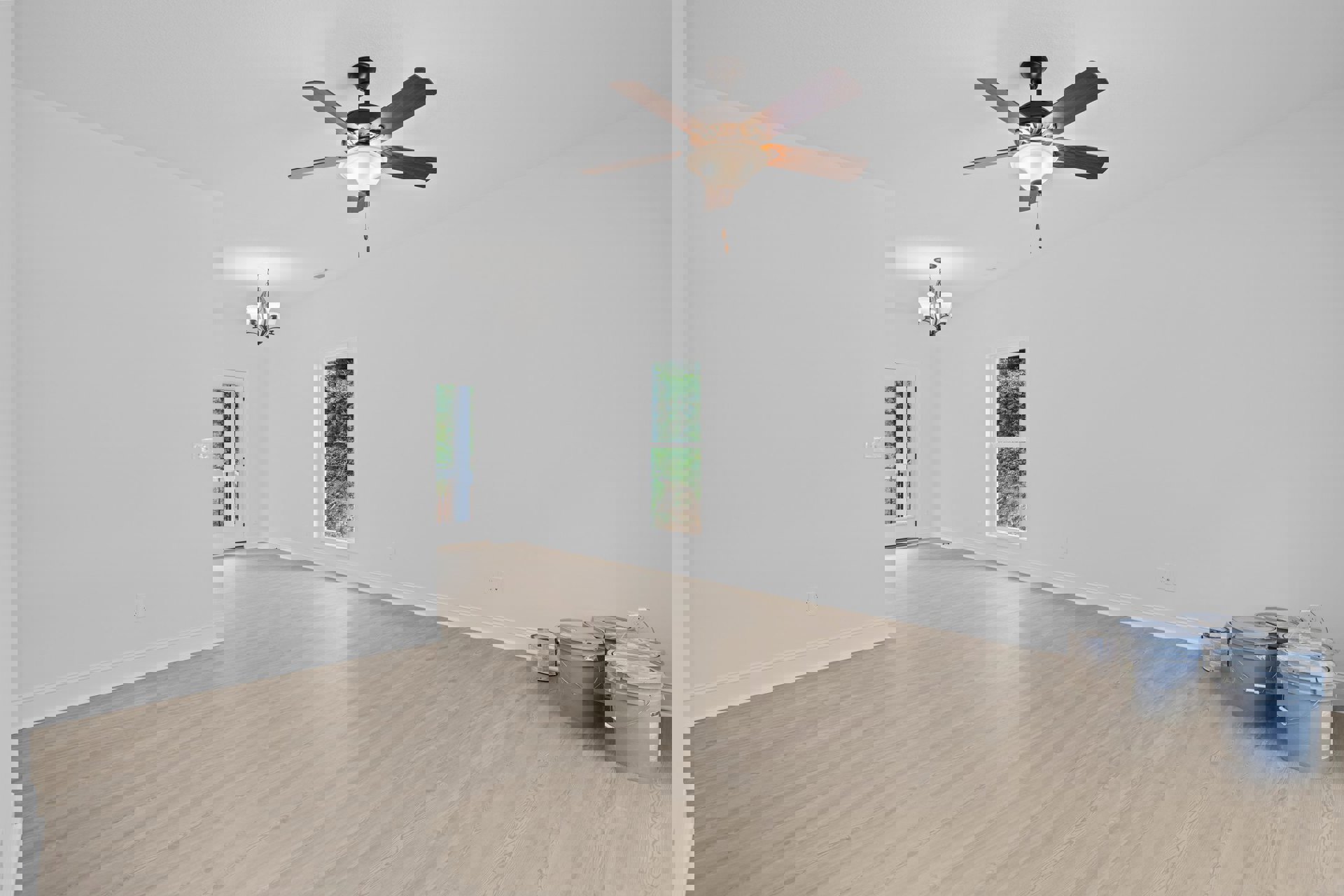 Ceiling fan with light fixture above wood laminate flooring, white-framed window, paint buckets and supplies on floor, neutral plaster walls