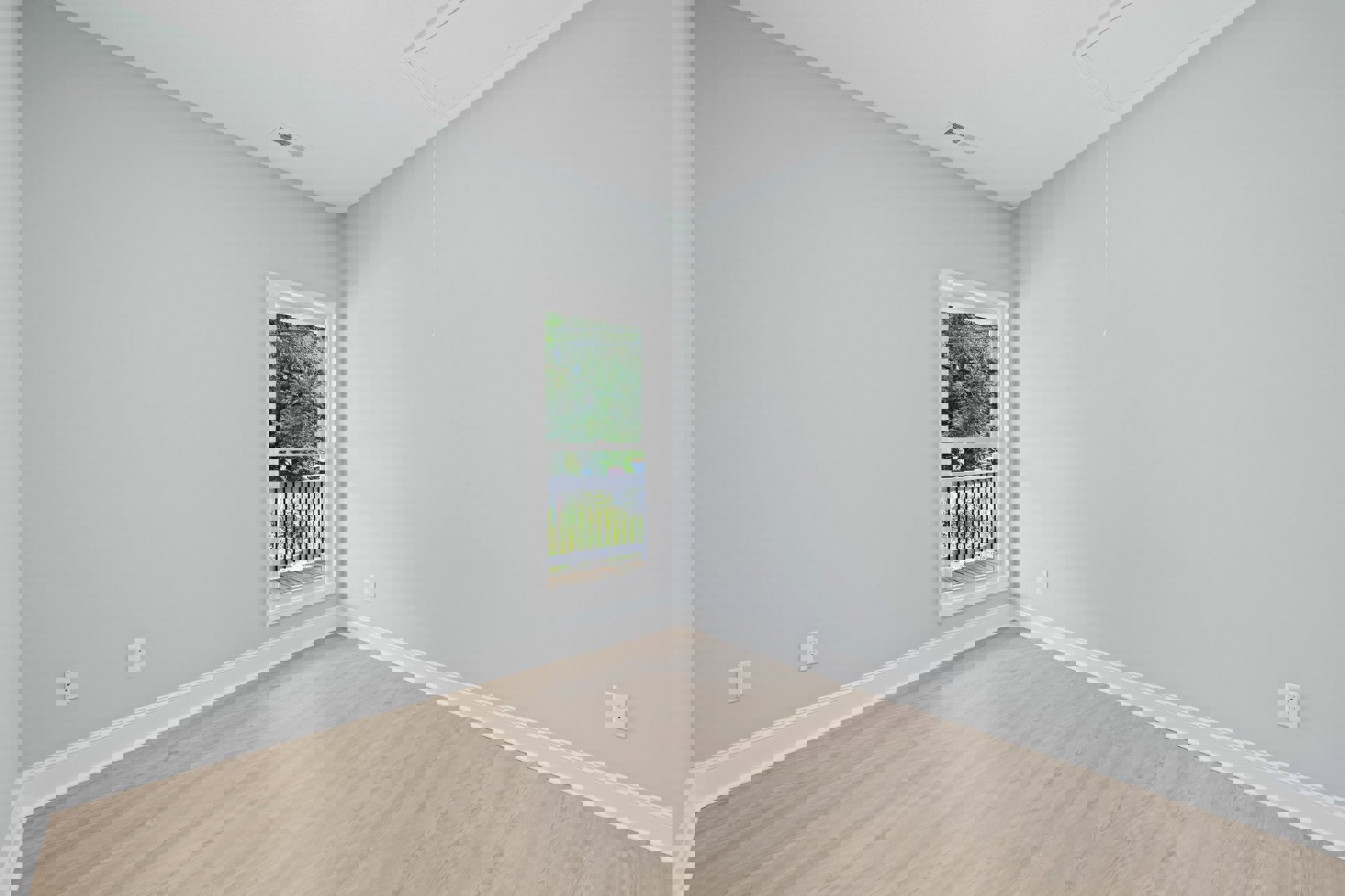 Sunlit room with wide window overlooking leafy trees, wood plank flooring, white plaster walls, balcony with wood decking and metal railing, double electrical outlet on wall