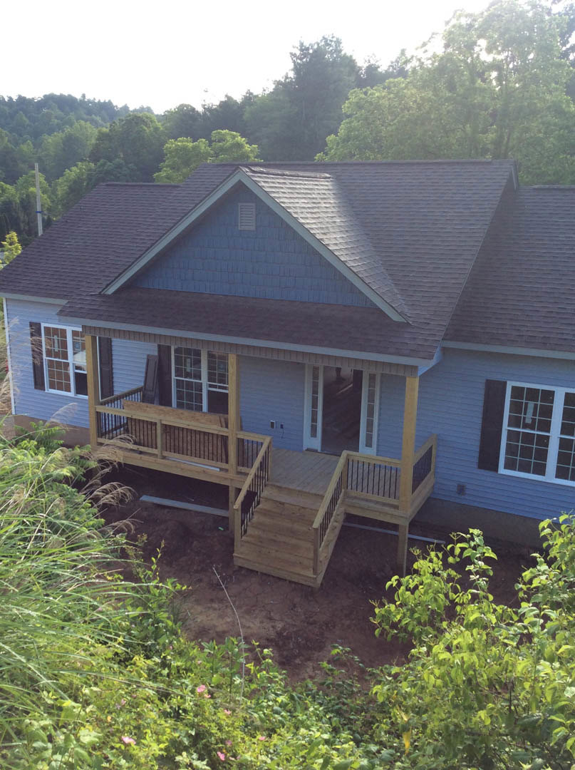 Two-story house with white-framed windows, wooden deck featuring metal railings and stairs, surrounded by mature trees and landscaped bushes.