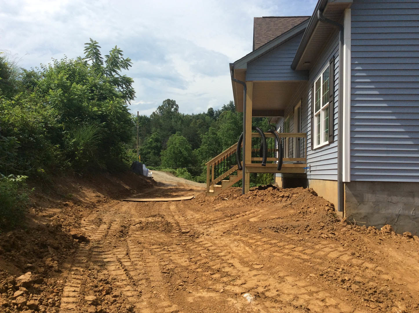 Dirt road with tire tracks leading to a modern house partially visible behind trees and bushes, wooden stairs with black railing, cloudy sky overhead, person in black jacket near