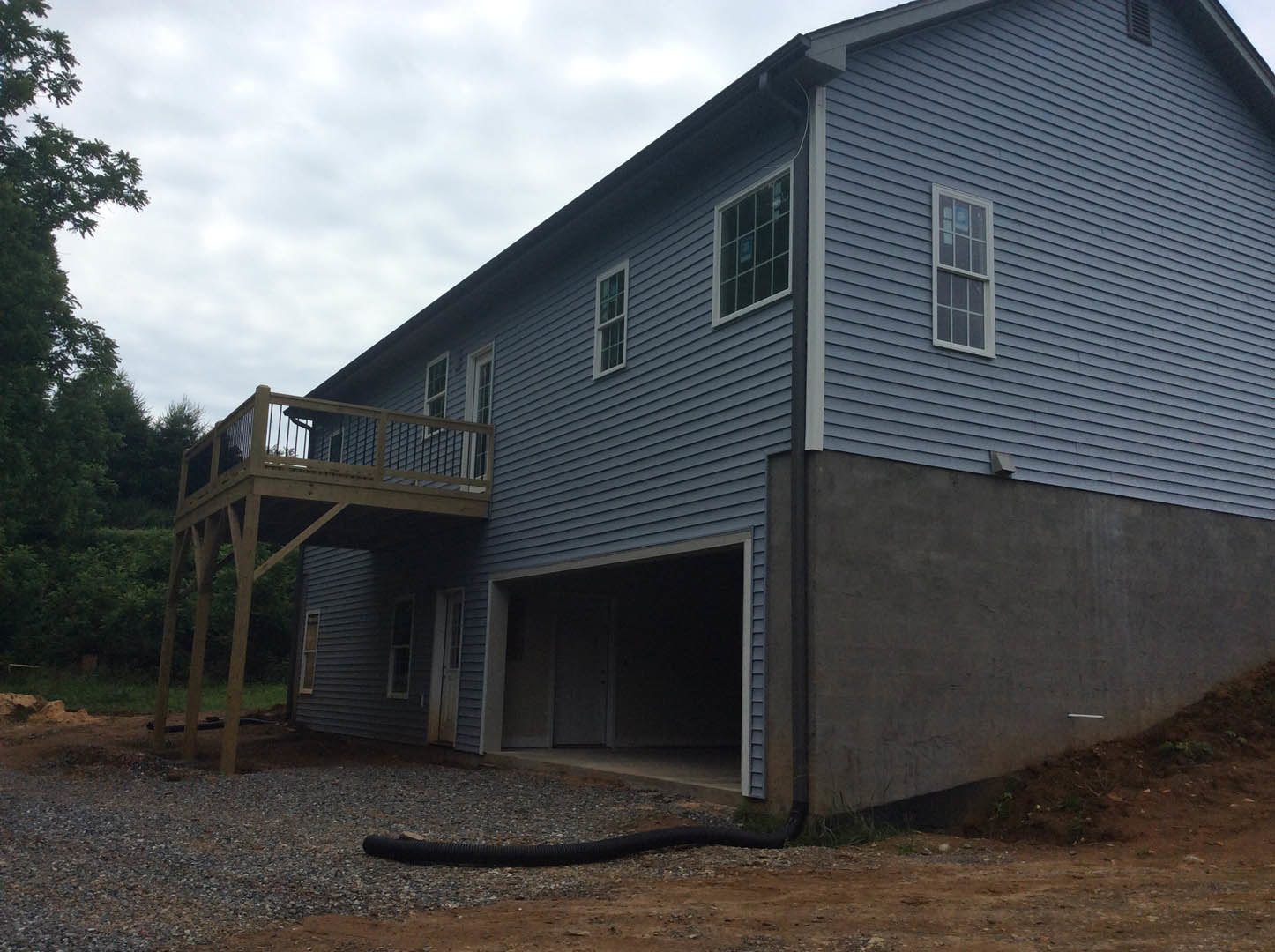 Two-story house with attached garage, elevated wooden deck, multiple square windows, light siding, and landscaped yard with trees under partly cloudy sky