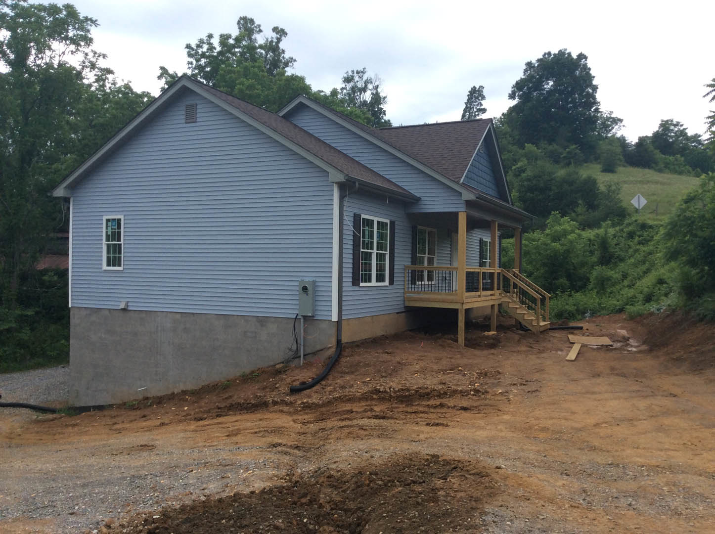Two-story house with multi-pane windows, wooden porch and deck featuring metal railing, dirt path leading to entry, surrounded by trees and plants under a clear sky.