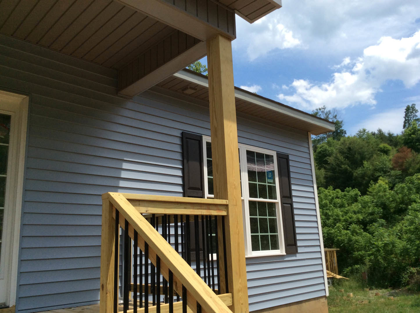 Front porch with wooden railing featuring black metal balusters, light-colored siding, large window, and stairs leading to entry, surrounded by trees and blue sky.