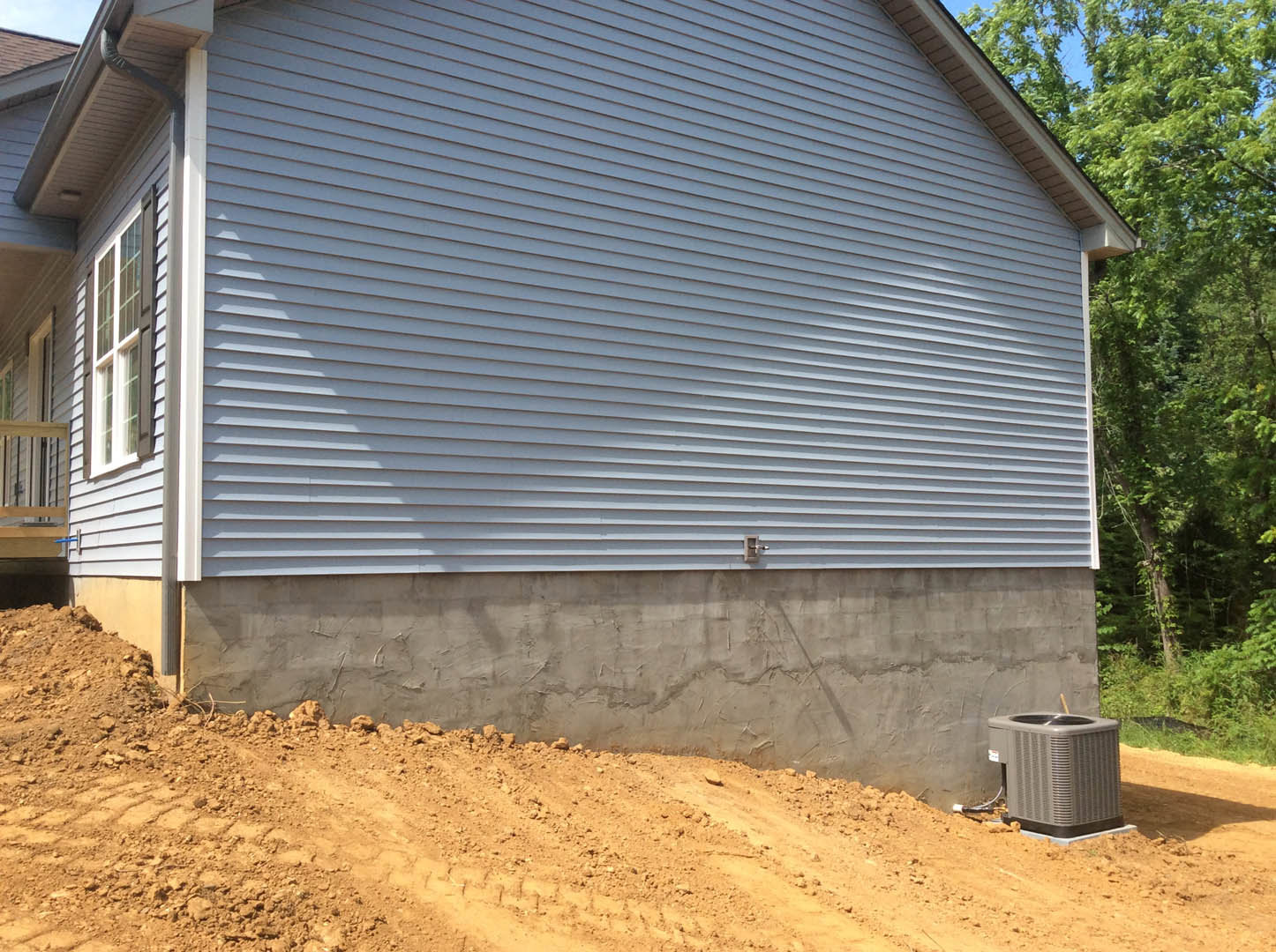 Modern home exterior with light grey siding, large windows, and a sloped dirt embankment alongside concrete foundation; small tree and plants visible near property edge.