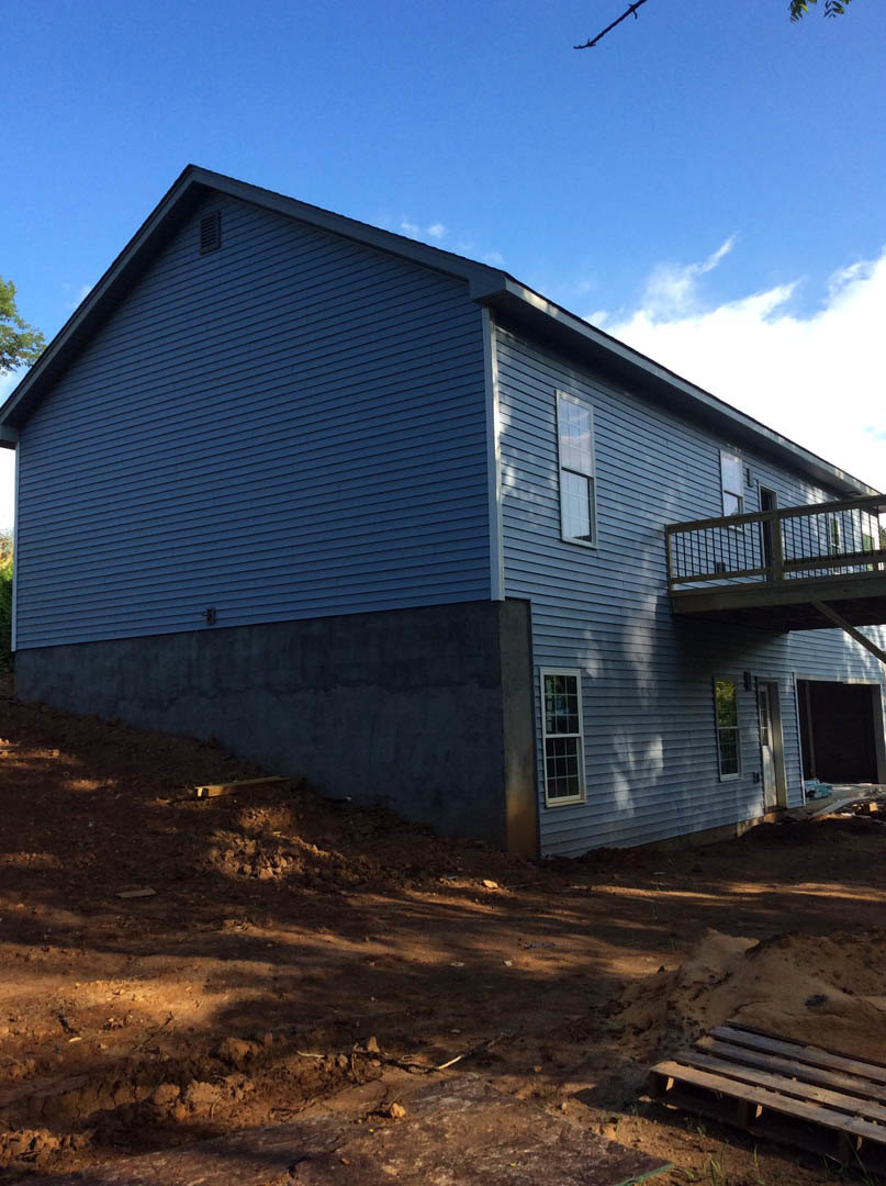 Two-story home under construction with exposed wooden deck, white siding, black front door, multi-pane windows, and scattered wooden pallets on dirt ground