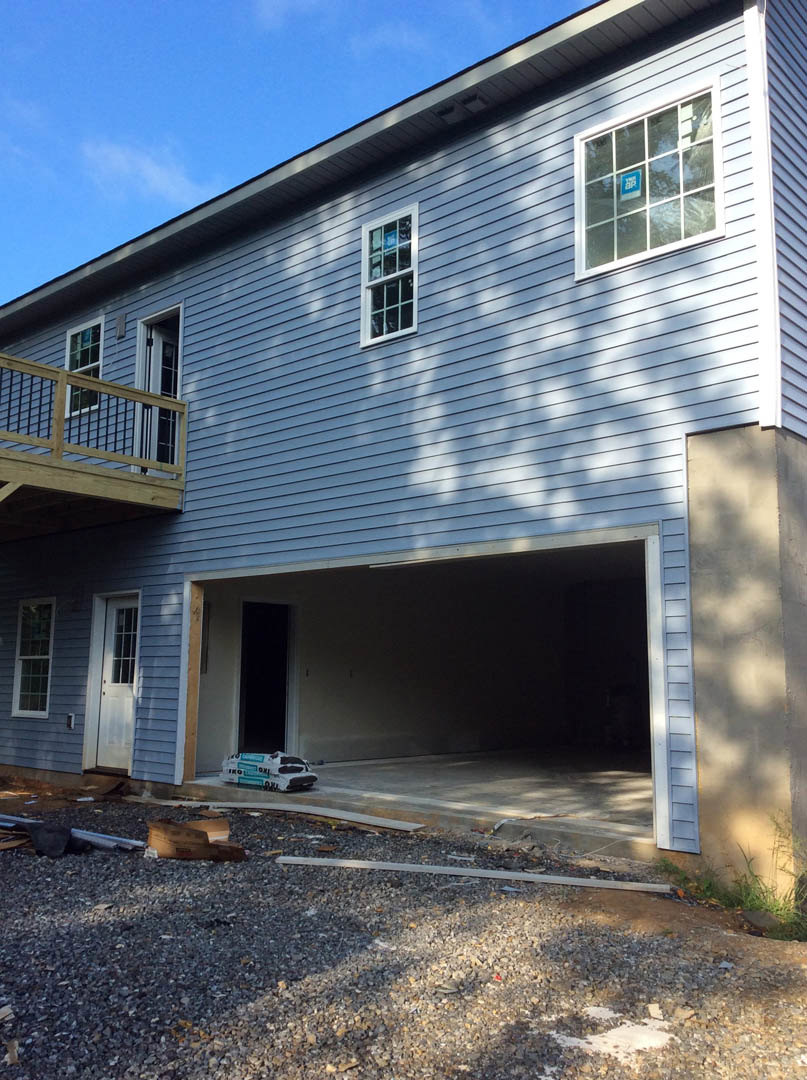 Partially built house with exposed wooden framing, attached garage, multi-pane windows, white door with glass insert, and unfinished deck under cloudy sky