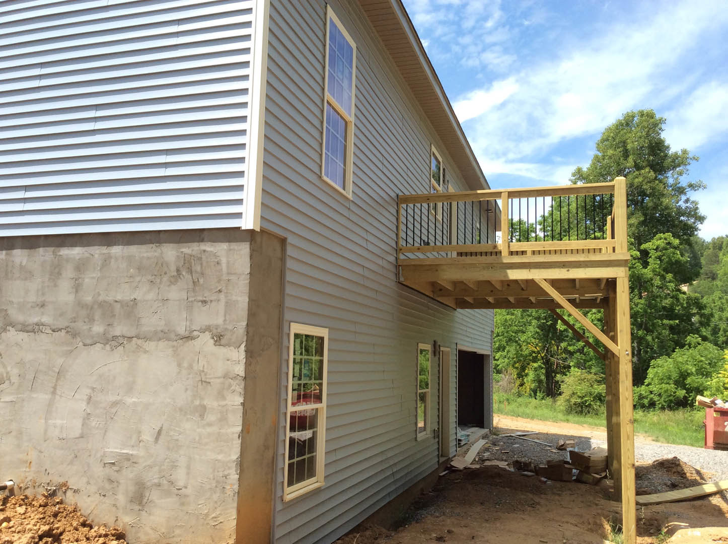 Modern home exterior featuring a wood deck, large windows, concrete retaining wall, and landscaped yard with brown soil and greenery.
