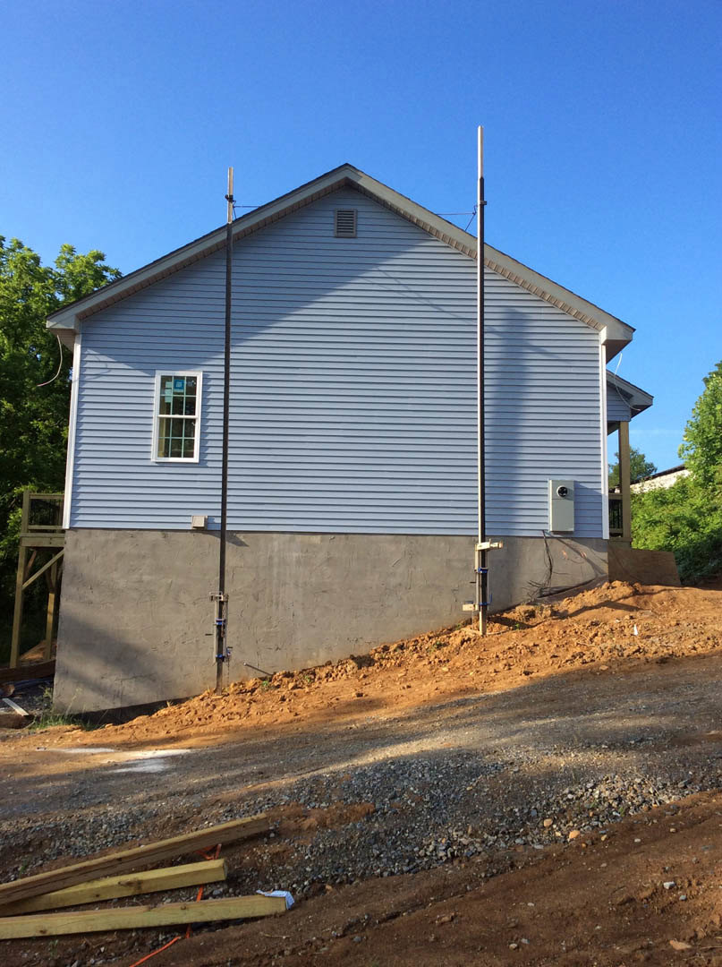 Modern home with exposed concrete foundation and wall, wooden deck, large window displaying a sign, pile of wood beside dirt road, small wooden shed, mature tree in background