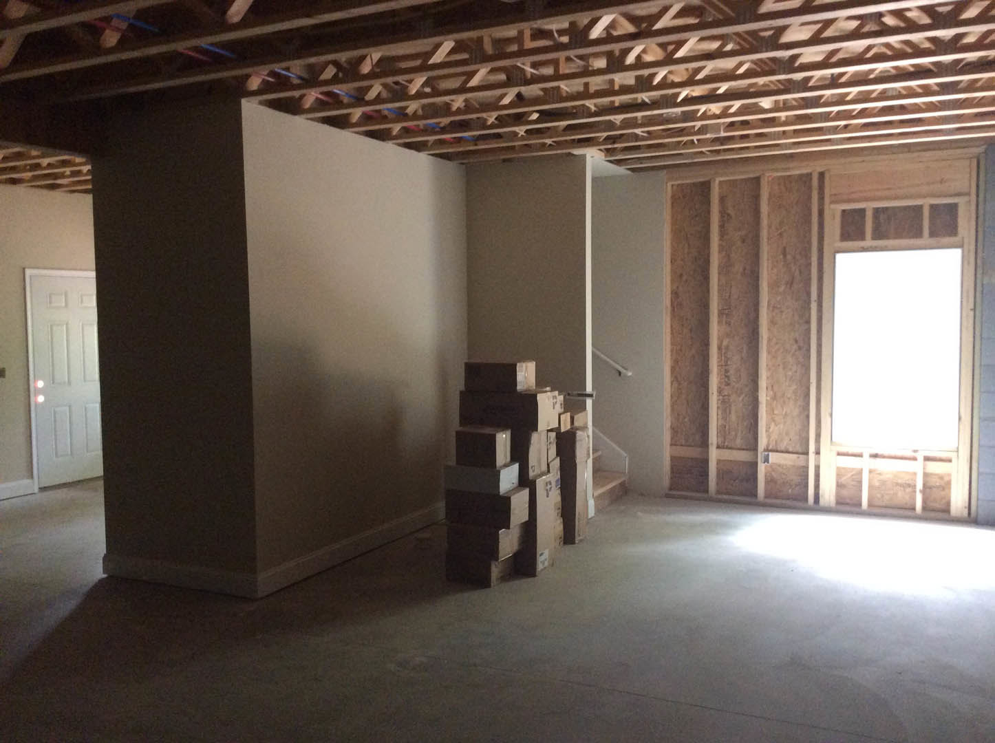 Unpacked boxes stacked on light wood flooring, white paneled door with overhead lighting, white plaster walls, window with trim, exposed wooden framing with drilled holes along one