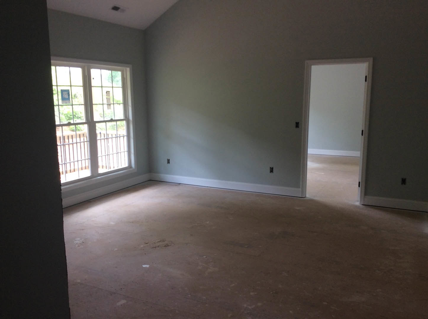 Spacious room featuring a white plaster wall, large window with metal fence, single door, and unfinished flooring.