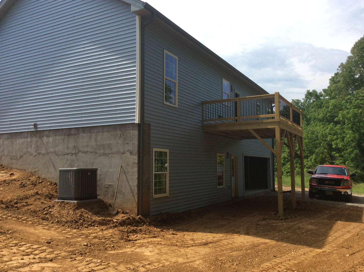 Two-story house with attached garage, red truck parked on gravel driveway, white-framed windows, wooden deck, surrounded by trees under partly cloudy sky