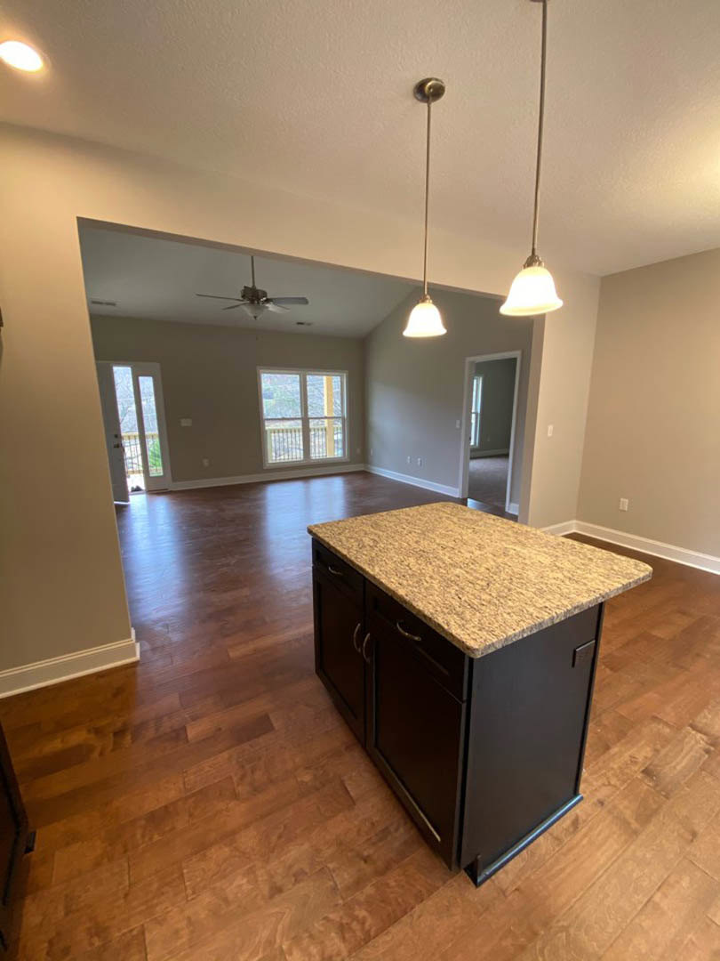 Marble-topped kitchen island with white cabinetry, laminate flooring, ceiling fan with light, large window featuring a metal railing, and smooth white walls.