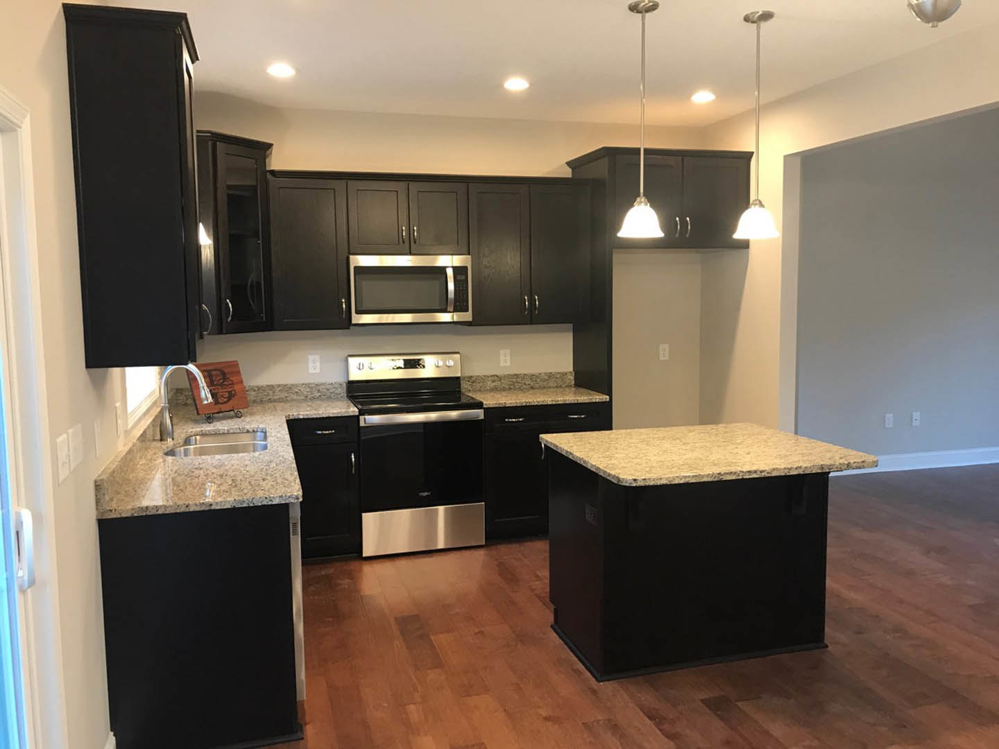 Black kitchen cabinets with granite countertops, stainless steel microwave and stove, black and white countertop, wooden sign with letter, light-colored flooring, modern fixtures.