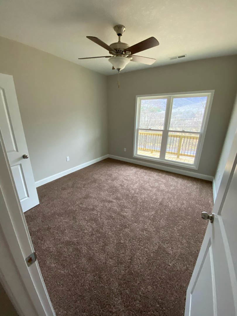 Ceiling fan with light fixture mounted on white plaster ceiling, large window with white railing, brown carpet flooring, neutral walls with crown molding