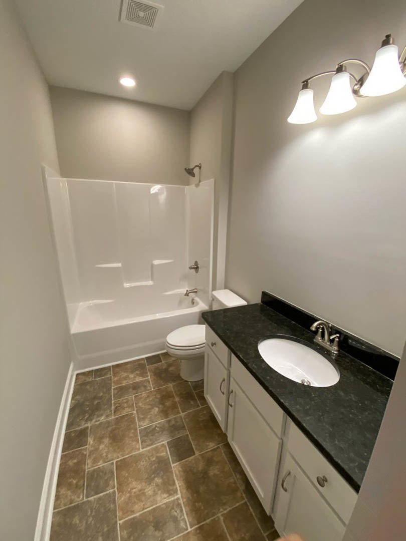 Modern bathroom with white porcelain sink, chrome faucet, toilet, glass-enclosed shower, light gray tile walls, dark wood vanity, and three-bulb overhead light fixture