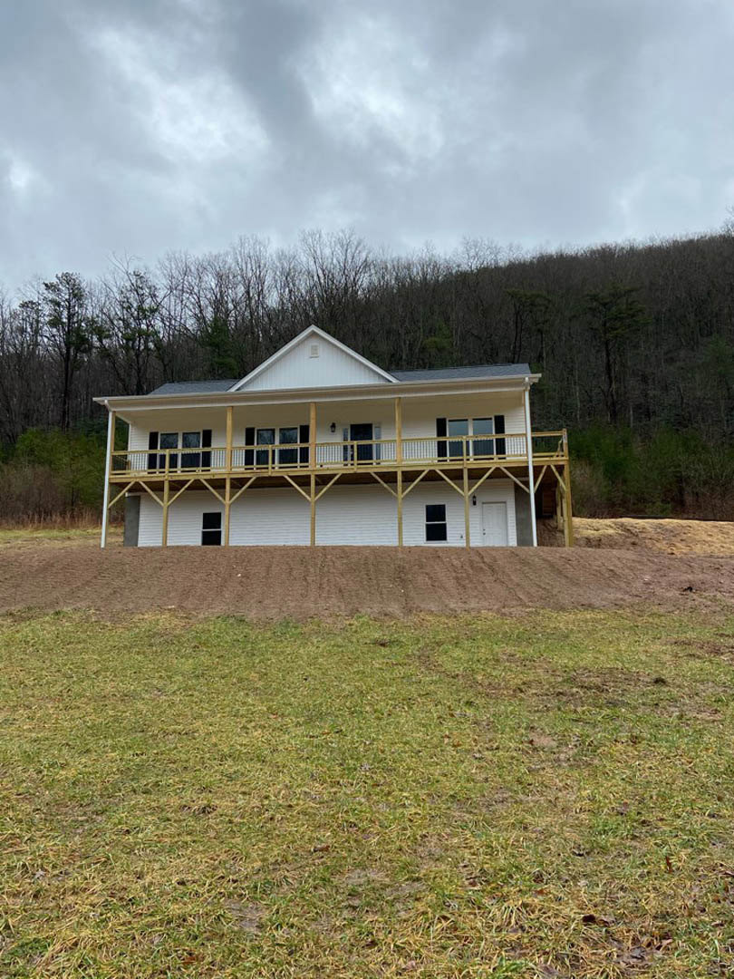 Two-story house with wide covered porch, white siding, balcony above, surrounded by grassy field, trees, and cloudy sky