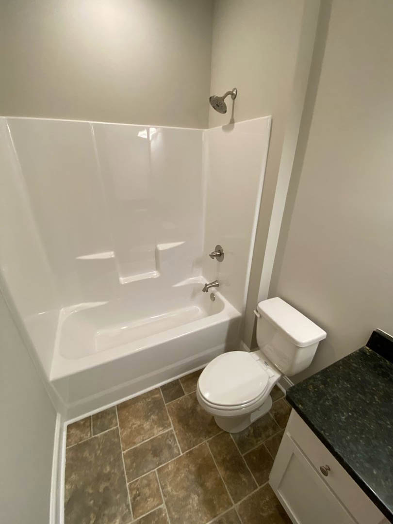 White bathtub and toilet beside a light-colored tile floor, with a close-up of a stone countertop and neutral wall finishes