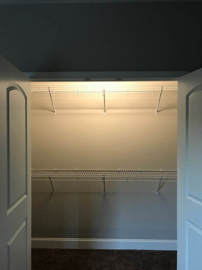 Walk-in closet with built-in white shelves, illuminated by recessed ceiling lights, featuring a metal hanging rod and grey carpeted floor.