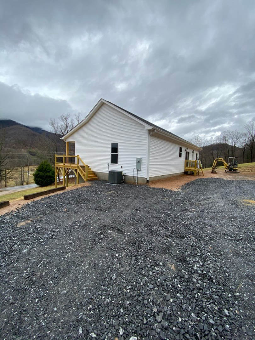 White house with yellow wooden staircase, gravel driveway in foreground, tree and grassy landscape surrounding exterior.