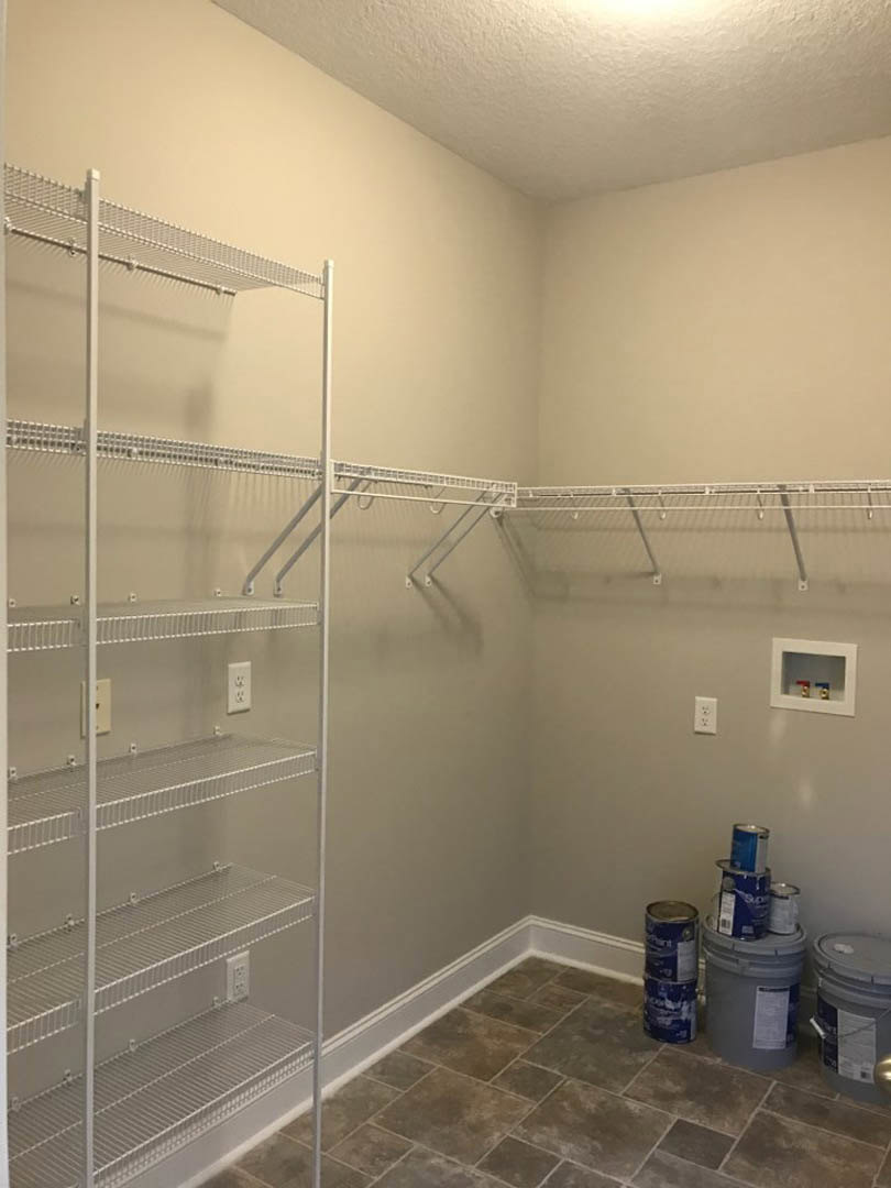White metal shelving stocked with paint cans against a white plaster wall, brown tile flooring with white baseboard, window with white frame, and grey storage container visible in