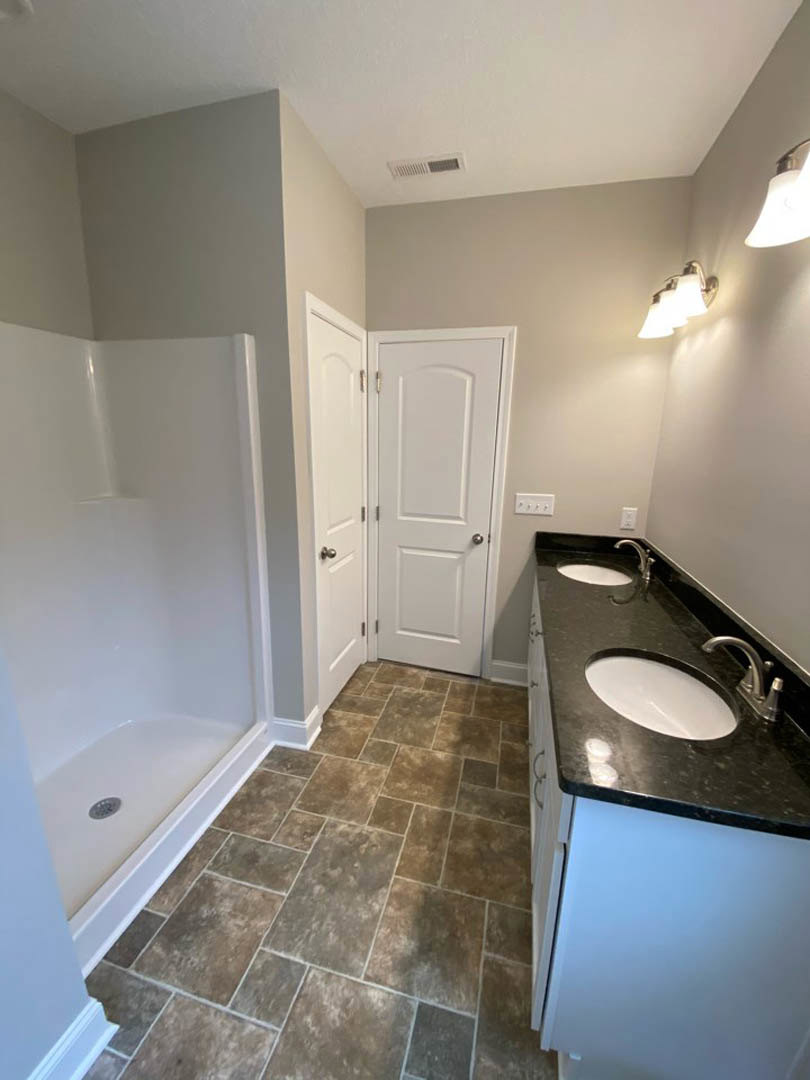 Double vanity bathroom with white sinks, quartz countertop, tile floor, glass-enclosed shower, white door with silver knob, and modern cabinetry.
