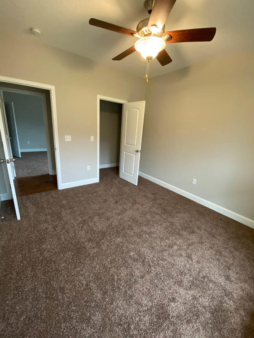 Carpeted room with white walls, ceiling fan with light fixture, and open white door with silver knob