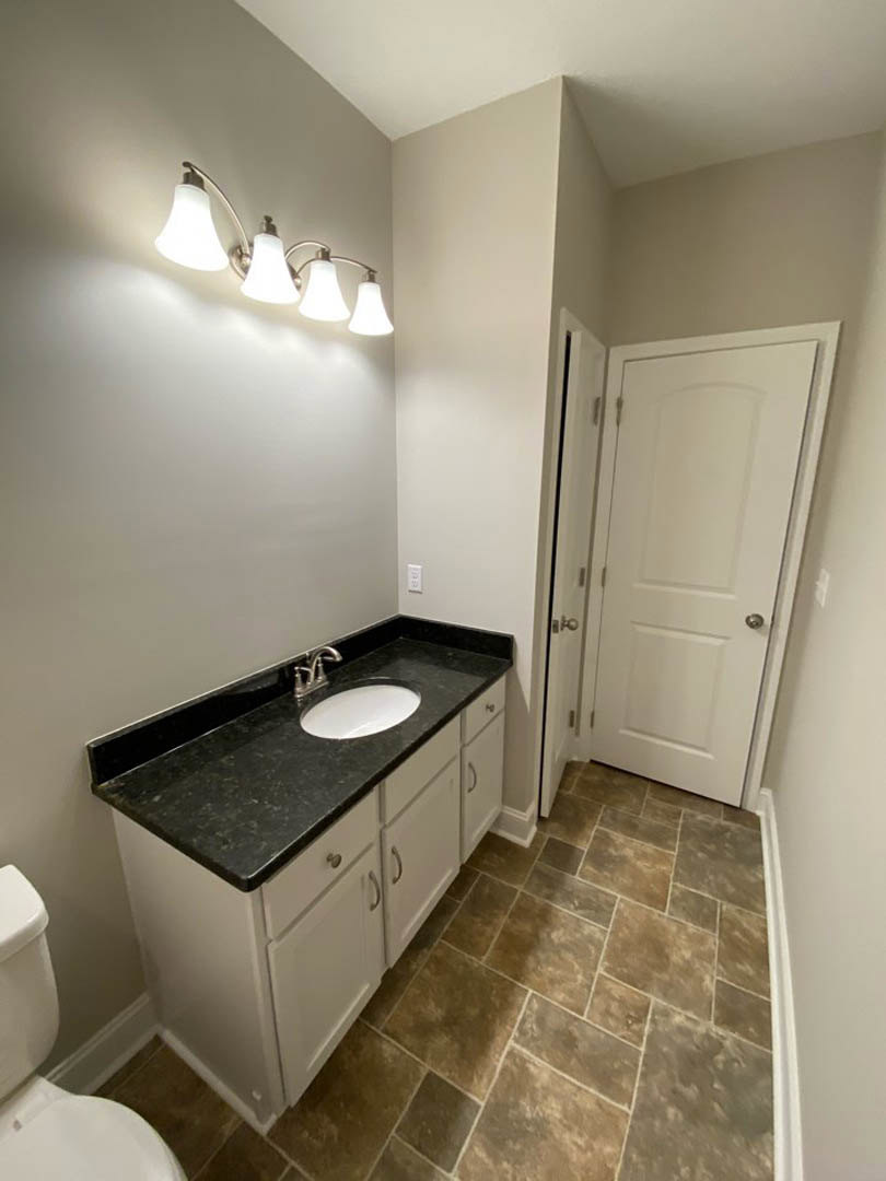 Bathroom with white porcelain sink and toilet, wall-mounted mirror above sink, chrome faucet, light fixture with four bulbs, tiled floor, white door with silver knob