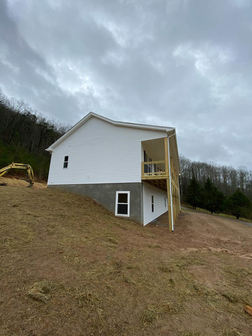Framed house under construction on a grassy hillside with exposed plywood, nearby yellow excavator arm, scattered trees, and cloudy sky