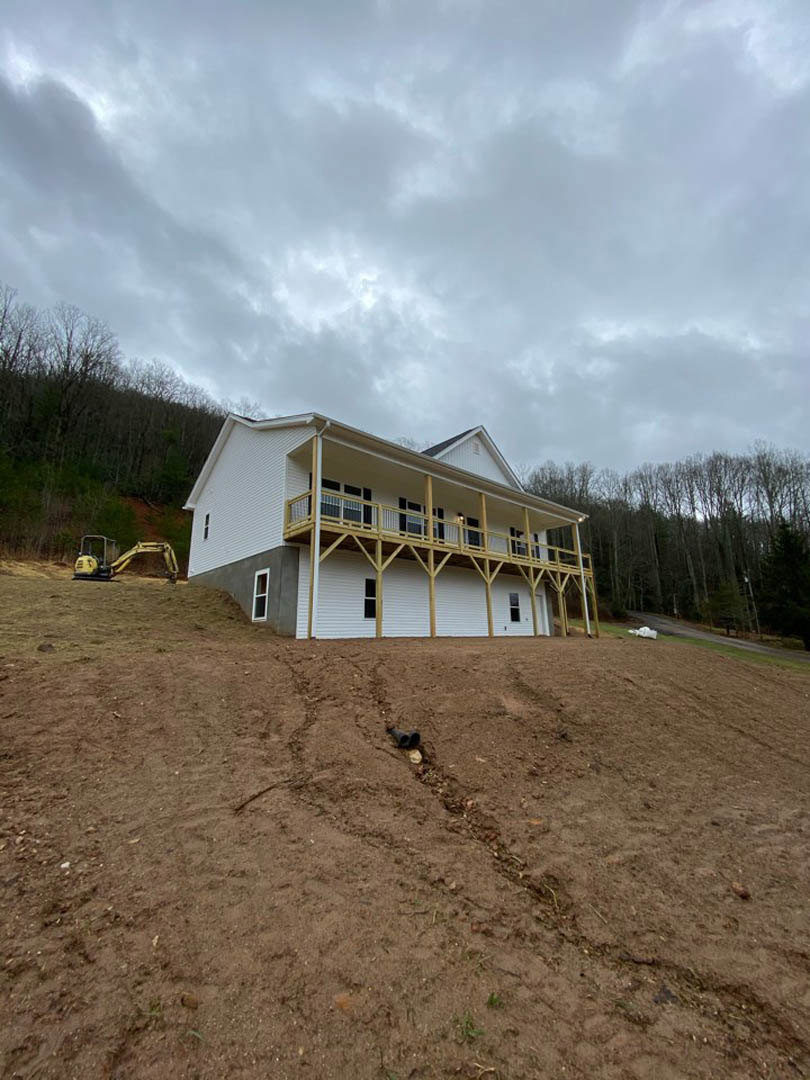 Two-story house with wide covered porch, light siding, and balcony; yellow bulldozer parked beside large dirt excavation; mature trees and cloudy sky in background