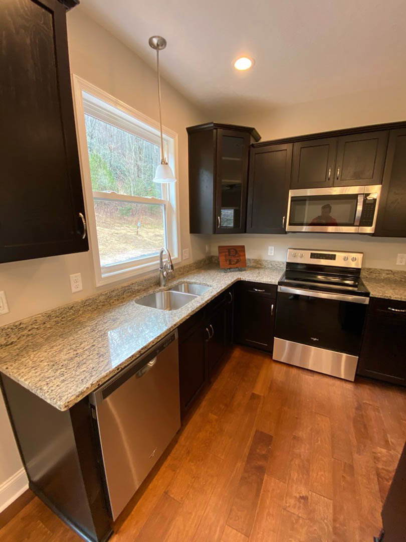 Granite countertop kitchen featuring stainless steel dishwasher, black and silver oven, white cabinetry, wooden sign with letter, window with natural light, and person in red shirt