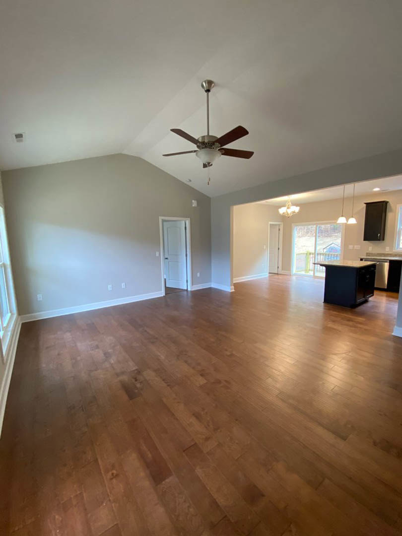 Spacious living room with hardwood flooring, ceiling fan featuring a light fixture, white walls, black rectangular fireplace with wooden mantel, and white door with black handle