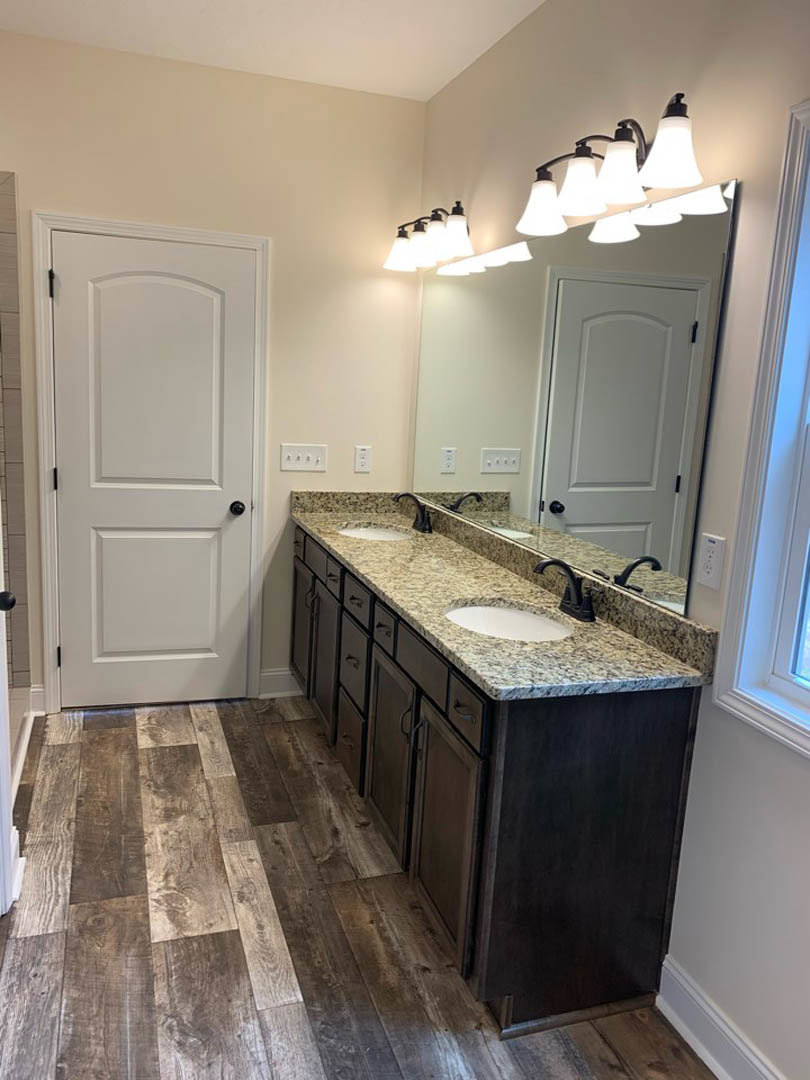 Bathroom with double sinks, wood flooring, white cabinetry, wall-mounted row of lights above mirrors, white door with black knob, and neutral tile accents